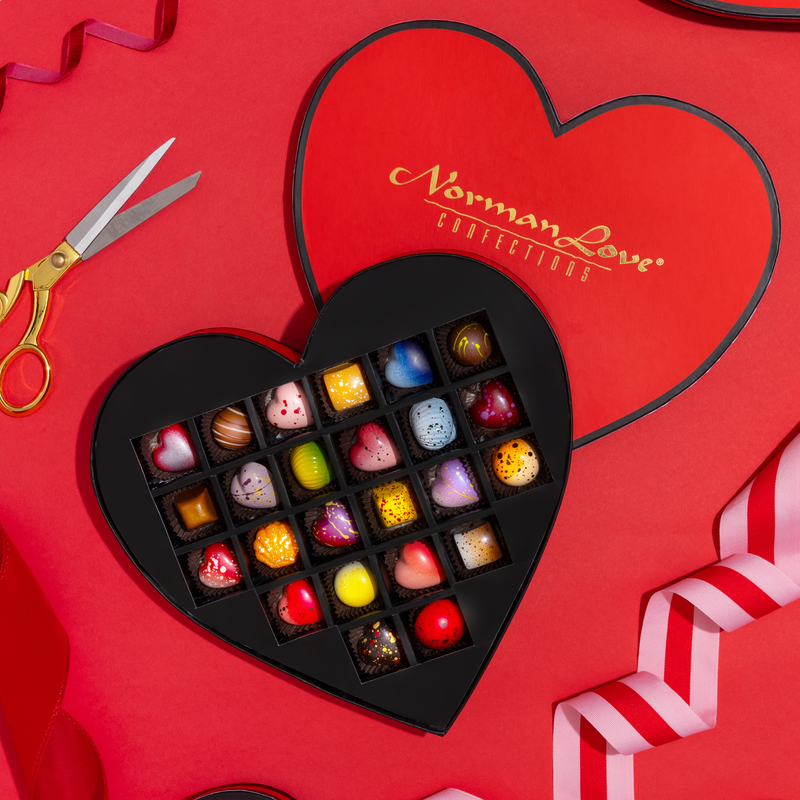 24 beautifully crafted chocolates in a red, heart-shaped gift box. The gift box includes 10 limited -edition hearts as well as 14 signature pieces. The box is displayed on a red background with a red ribbon and gold scissors.